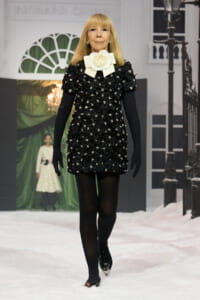 Model walking a wintery runway in a black floral-embellished mini dress with a large cream bow at the neck and long black gloves to the elbows.