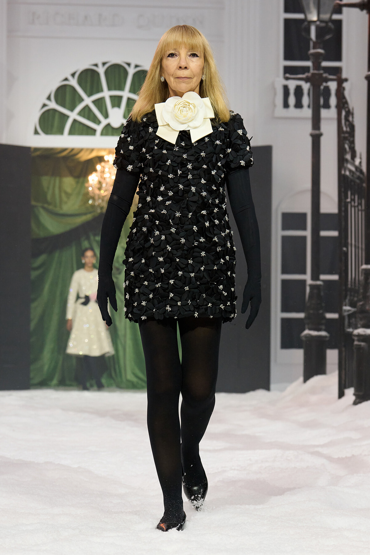 Model walking a wintery runway in a black floral-embellished mini dress with a large cream bow at the neck and long black gloves to the elbows.