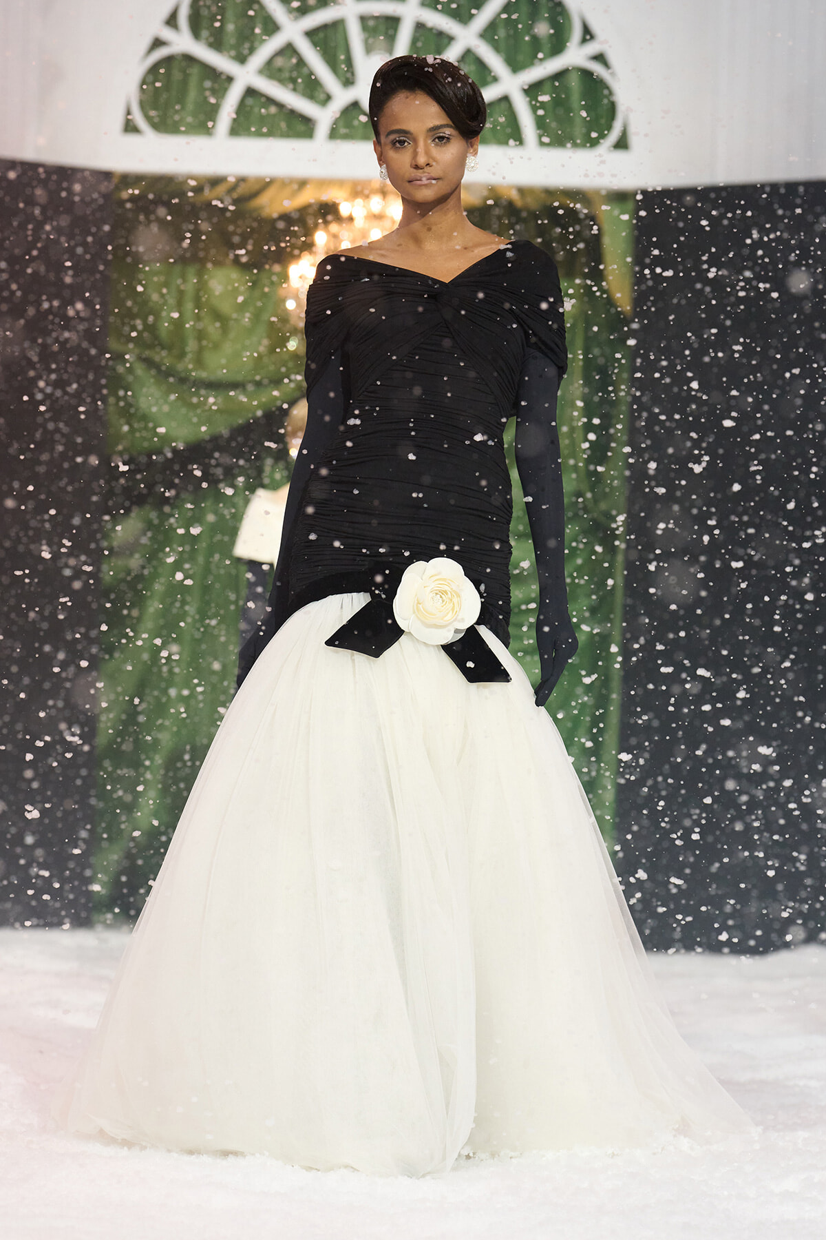 Model walking in a black off-shoulder top and white voluminous gown, snow falling around on a runway.