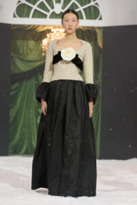 Model walks a runway in a two-tone gown: beaded ivory bodice with a large white flower and black bow, and a full black skirt; snow on the floor.