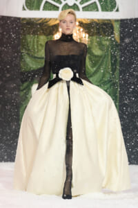 Fashion model in a black sheer bodice and a voluminous ivory ball gown with a white rose belt, standing on a snowy runway.