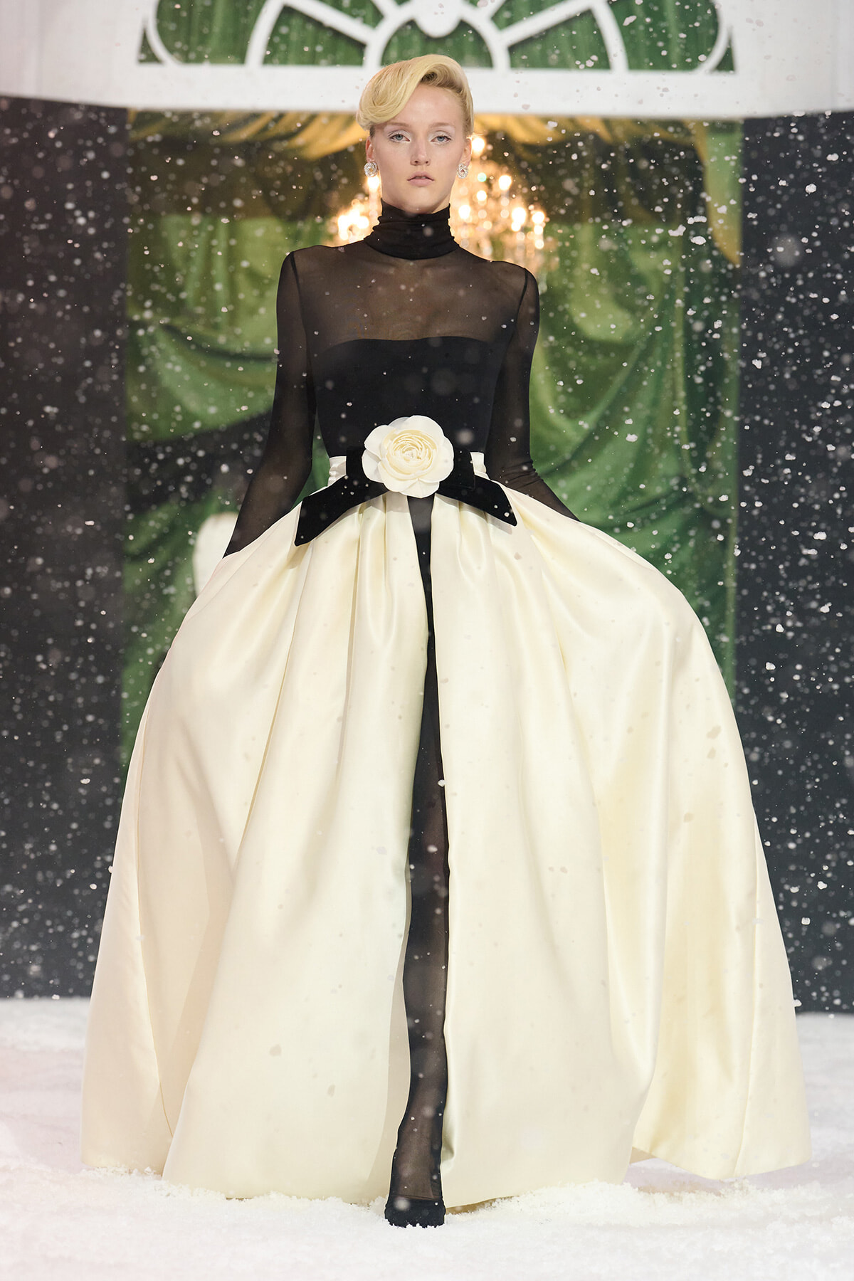 Fashion model in a black sheer bodice and a voluminous ivory ball gown with a white rose belt, standing on a snowy runway.