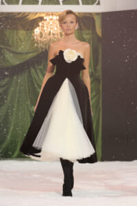 Model walking a runway in a black strapless ballgown with a white tulle insert and large white flower at the bodice, on a snowy stage backdrop.
