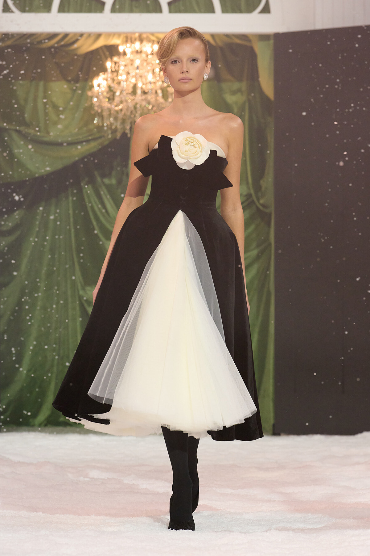 Model walking a runway in a black strapless ballgown with a white tulle insert and large white flower at the bodice, on a snowy stage backdrop.