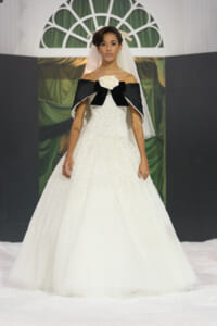 Model wearing an off-the-shoulder white beaded wedding gown with a black capelet, white rose bow, and veil on a runway