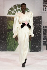 Model on a runway wearing an ivory satin gown with dramatic puff sleeves, a black bow belt, and a pink rose, walking through artificial snow.