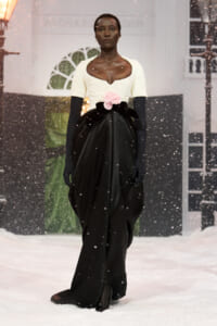 Model walking a snowy runway in a white lace bodice, a voluminous black skirt, pink rose belt, and long black gloves.
