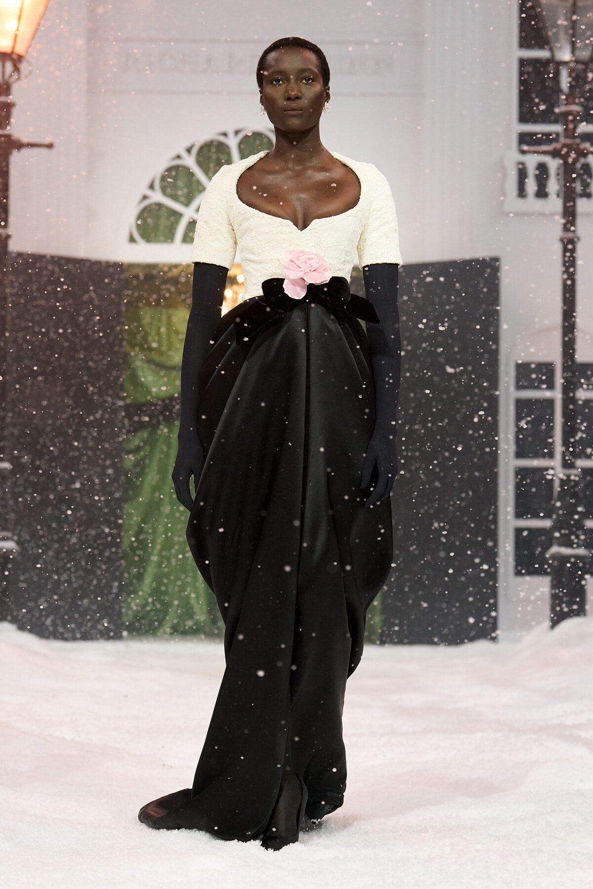 Model walking a snowy runway in a white lace bodice, a voluminous black skirt, pink rose belt, and long black gloves.