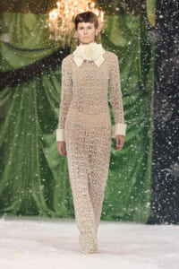Model walks a runway in a beige lace jumpsuit with an oversized cream bow collar and matching cuffs, snowflakes falling around a green backdrop.