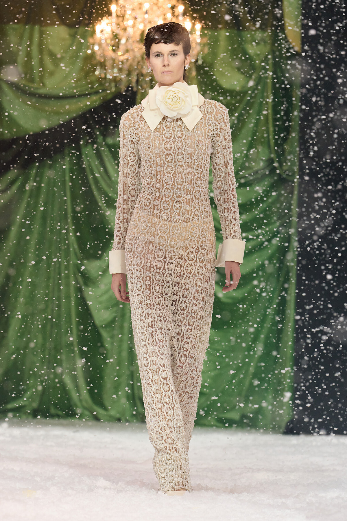 Model walks a runway in a beige lace jumpsuit with an oversized cream bow collar and matching cuffs, snowflakes falling around a green backdrop.