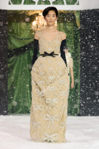 Model walking a snowy runway in a beige floor-length gown with silver bow embroidery, black off-the-shoulder sleeves, and a black belt.