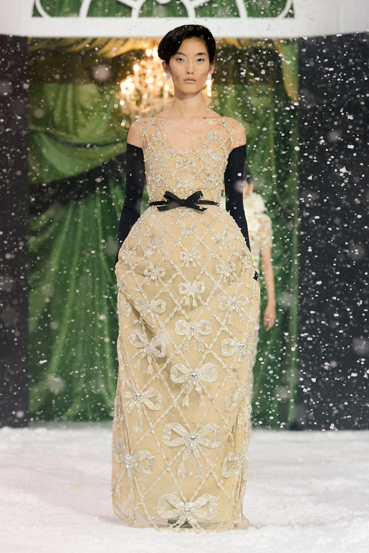 Model walking a snowy runway in a beige floor-length gown with silver bow embroidery, black off-the-shoulder sleeves, and a black belt.