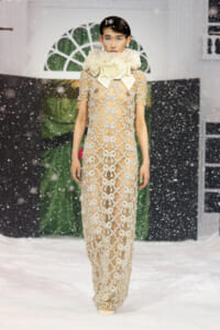 Model wearing a beige sheer gown with silver bow appliqués and a large white rose collar on a snowy runway show