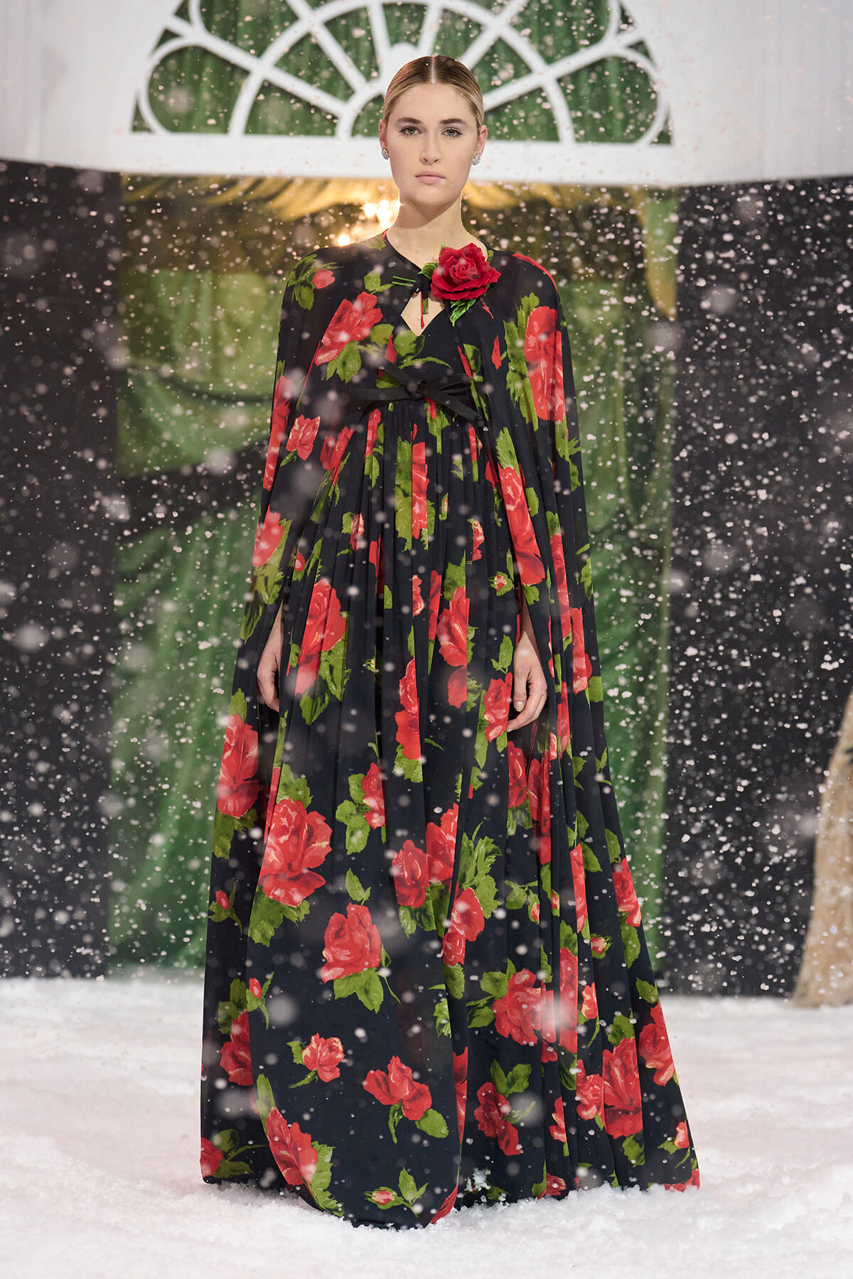 Model in a floor-length black gown with bold red rose print and green leaves, a red rose brooch at the neckline, standing on a snowy runway.