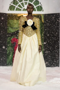 Model walks a winter runway in a gold sequin bodice and full cream skirt, adorned with a white rose and black bow at the chest as snow falls.