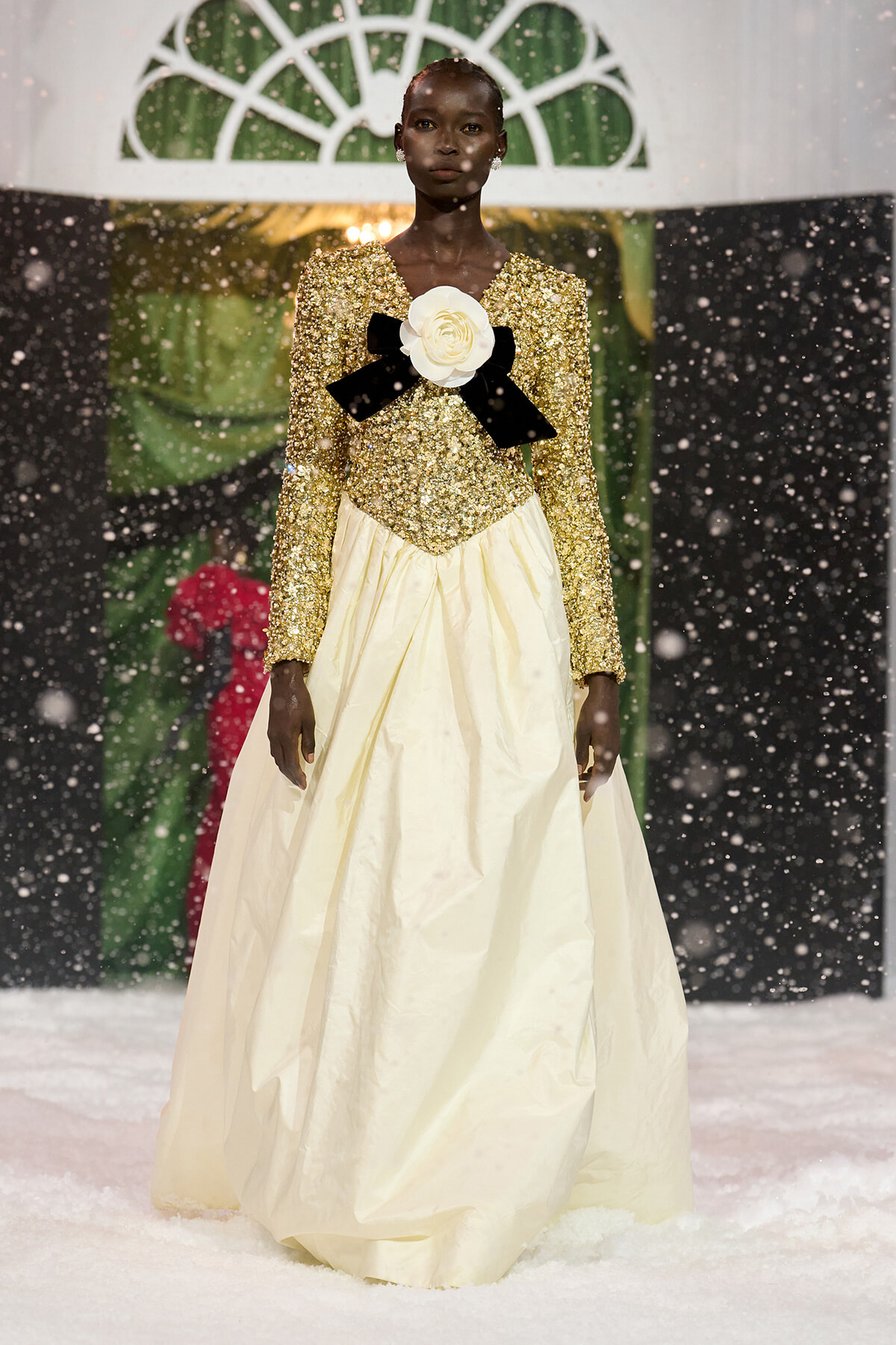 Model walks a winter runway in a gold sequin bodice and full cream skirt, adorned with a white rose and black bow at the chest as snow falls.