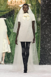 Model on a snowy runway wearing a cream pearl-beaded mini dress with a large cream bow at the neck, and a sheer veil and headpiece.