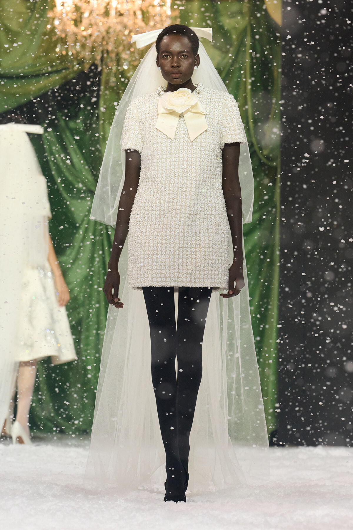 Model on a snowy runway wearing a cream pearl-beaded mini dress with a large cream bow at the neck, and a sheer veil and headpiece.