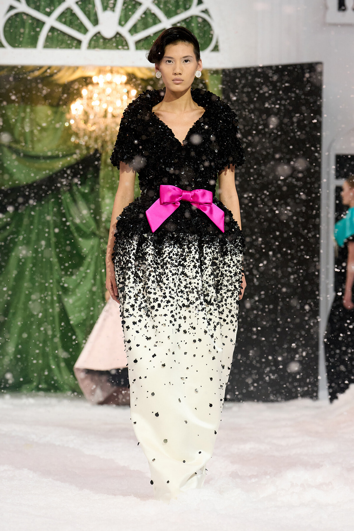 Model walking a snowy runway in a black textured top with a bright pink bow at the waist and a white, speckled gradient skirt.
