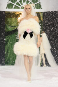 Model walks a snowy runway in an ivory feathered bustier dress with a pink flower and black ribbon belt, paired with a veil and headpiece, snowfall around.