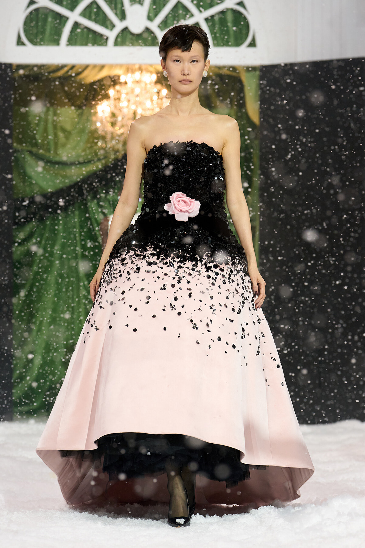 Model walks the runway in a strapless black-to-pink gradient ball gown with a pink rose at the waist, snow-like confetti falling around her.