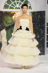 Model on a snowy runway wearing a strapless cream ball gown with tiered ruffles, a black waist belt with a white flower, and a veil.”] ,