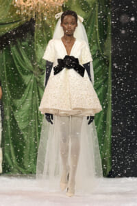 Model in an ivory embellished dress with a large black bow at the waist, veil, and white gloves walking on a snowy runway.