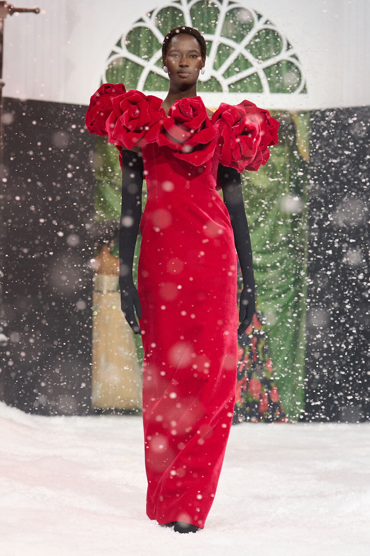 Model walking in a red gown with dramatic ruffle shoulders on a snowy runway. The bold dress contrasts with black sleeves and white snowfall.