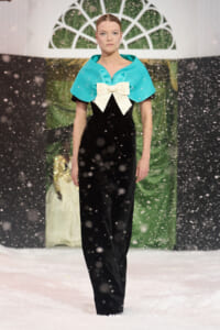 Model walks a snowy runway wearing a turquoise capelet with a cream bow over a black velvet jumpsuit.