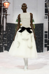 African model walks a white beaded evening gown with a high neckline and a large black bow at the waist, snow falling on a fashion runway.