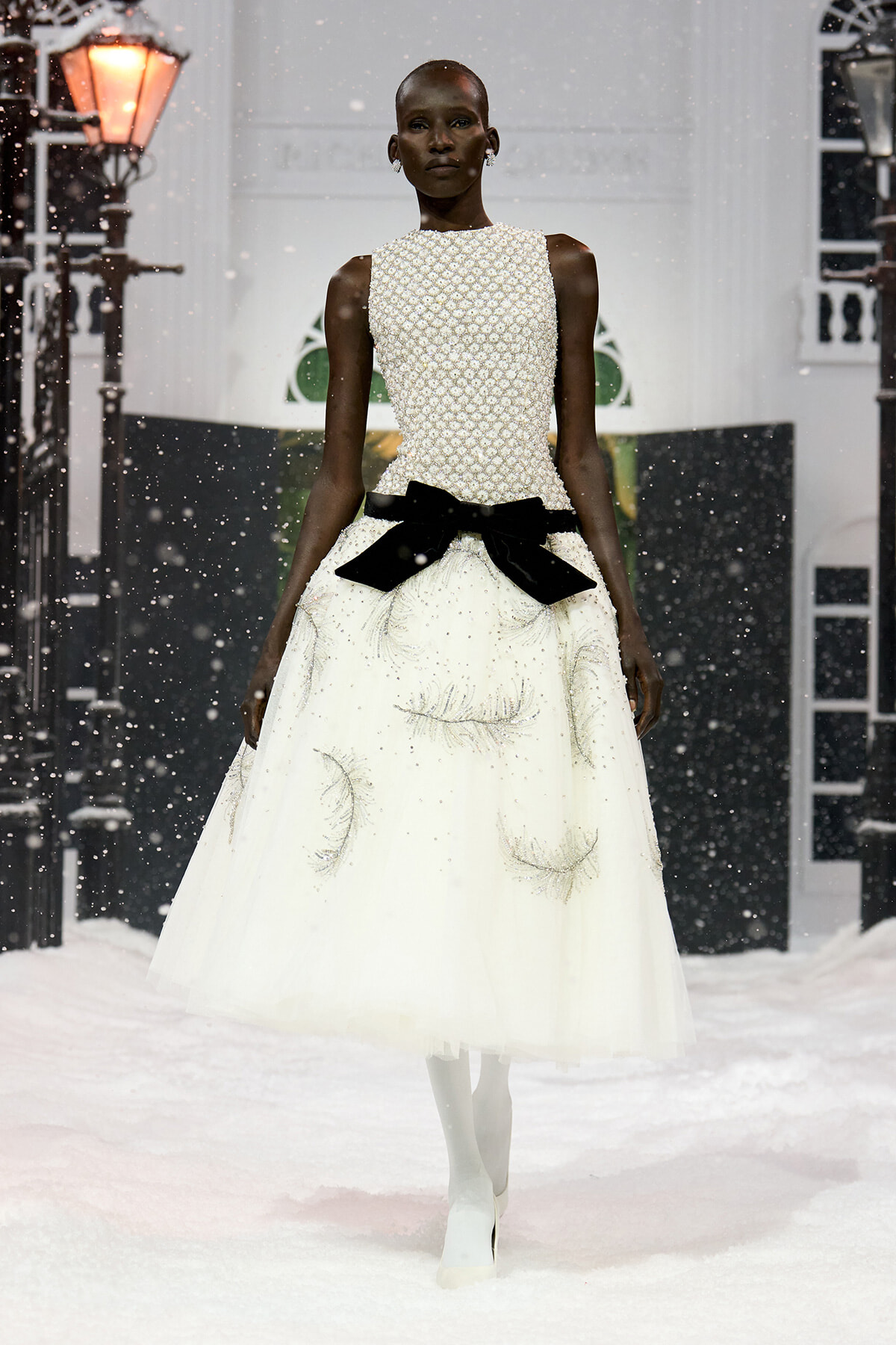 African model walks a white beaded evening gown with a high neckline and a large black bow at the waist, snow falling on a fashion runway.