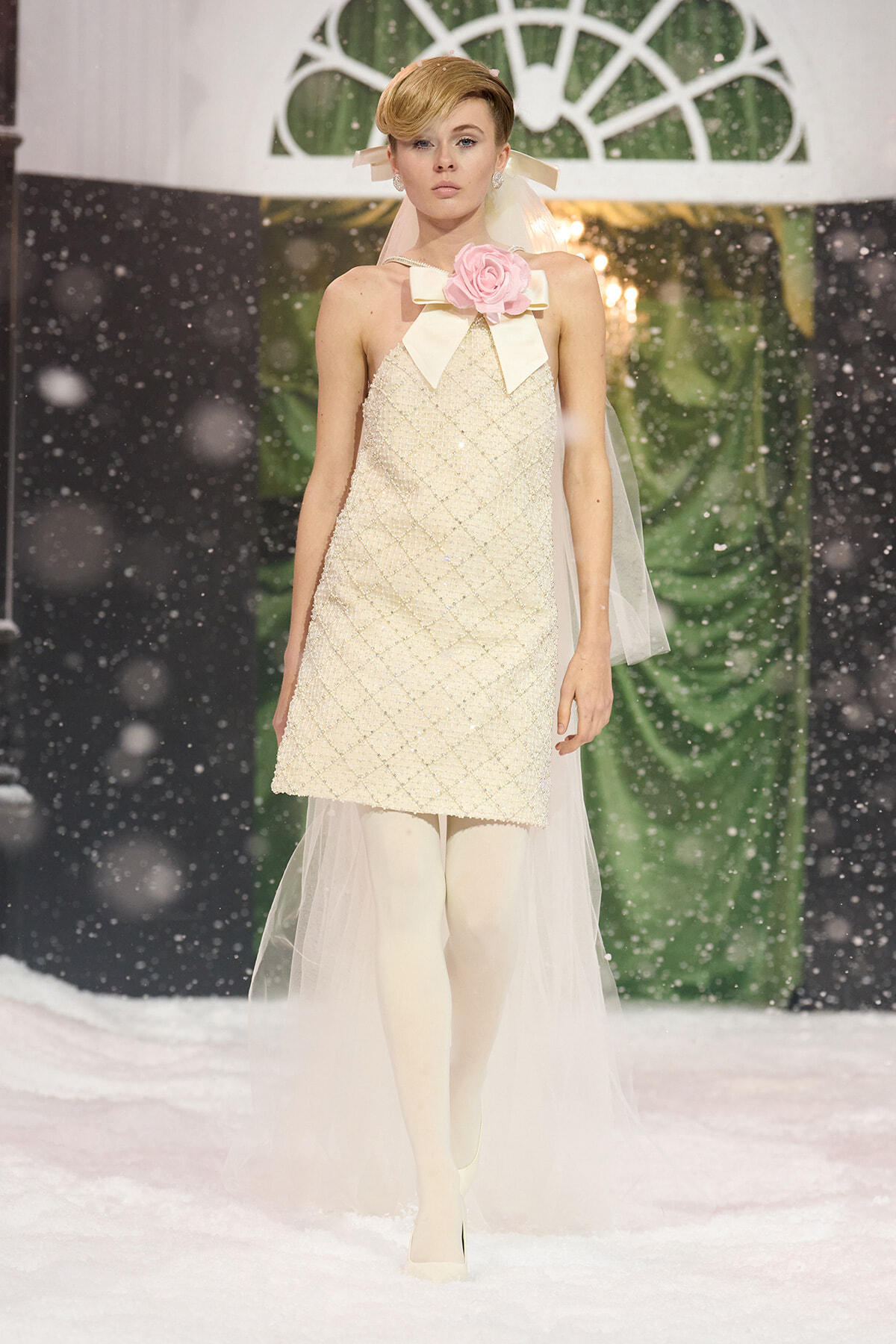 Model wearing a cream beaded shift dress with a pink rose bow at the neckline, veil, and white tights on a snowy runway.