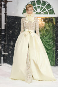 Model wearing an ivory lace ball gown with a high ruffled collar and oversized bow at the waist, standing on a snowy runway.