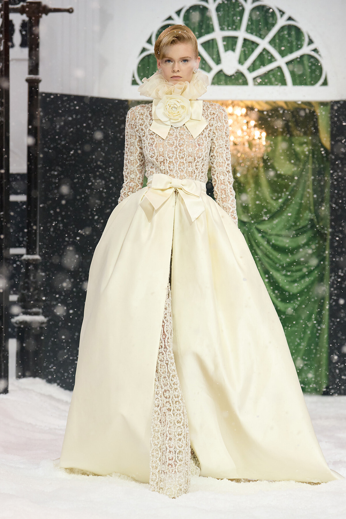 Model wearing an ivory lace ball gown with a high ruffled collar and oversized bow at the waist, standing on a snowy runway.