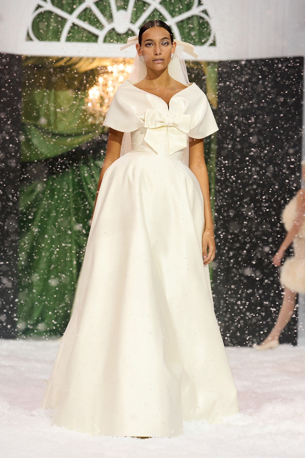 Model wearing a white bridal gown with a large bow at the waist on a snowy fashion runway.