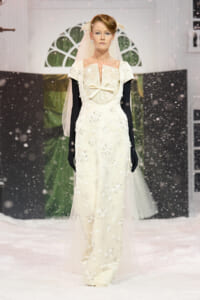 Model walks a snowy runway in an ivory lace wedding dress with a sheer cape, veil, and long black gloves.