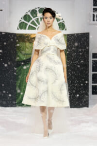 Model walks a snowy runway in a white off-the-shoulder dress with silver fern embroidery and veil in place