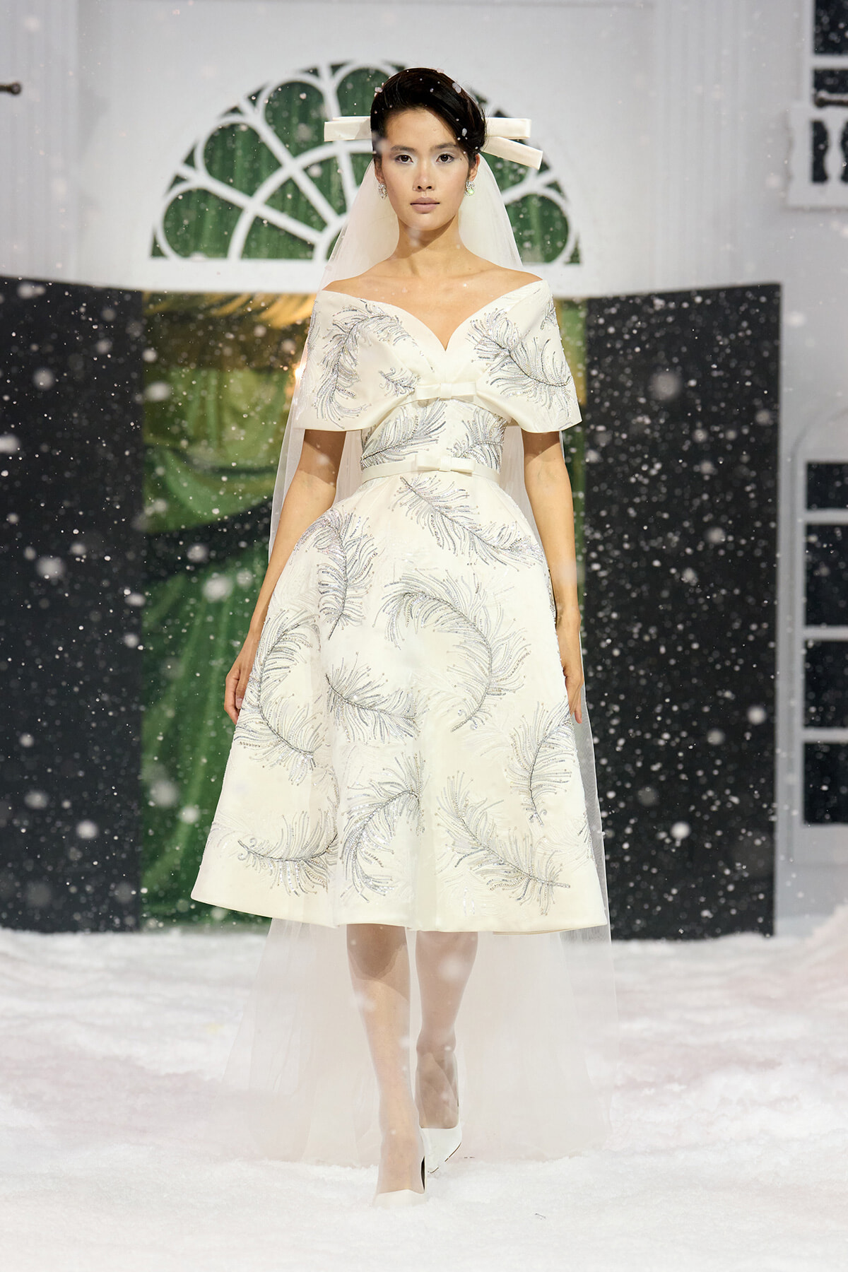 Model walks a snowy runway in a white off-the-shoulder dress with silver fern embroidery and veil in place