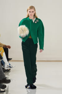 Model walking a runway in a bright green knit cardigan with brooches, dark green pants, and a fluffy cream handbag.