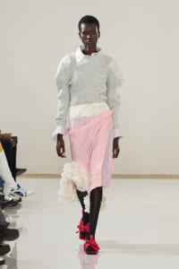 Model walking runway in a light gray oversized knit sweater layered over a white checkered shirt, paired with a pastel pink and cream wrap skirt featuring ruffled cream detail at the hem and bright pink bow-tied sandals.