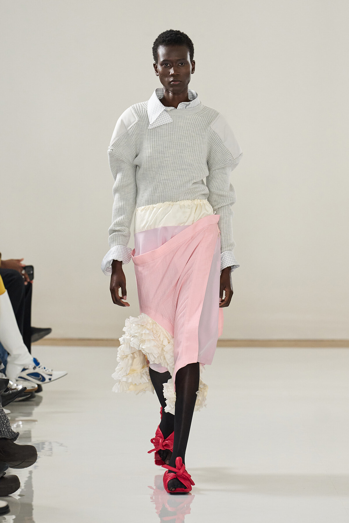 Model walking runway in a light gray oversized knit sweater layered over a white checkered shirt, paired with a pastel pink and cream wrap skirt featuring ruffled cream detail at the hem and bright pink bow-tied sandals.