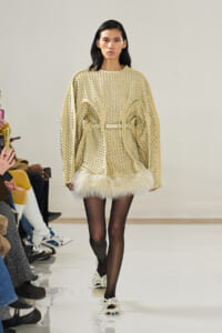 Model walking a runway in a beige oversized studded sweater with a belt, and white fur trim at the hem, paired with black tights and fur-accented shoes.