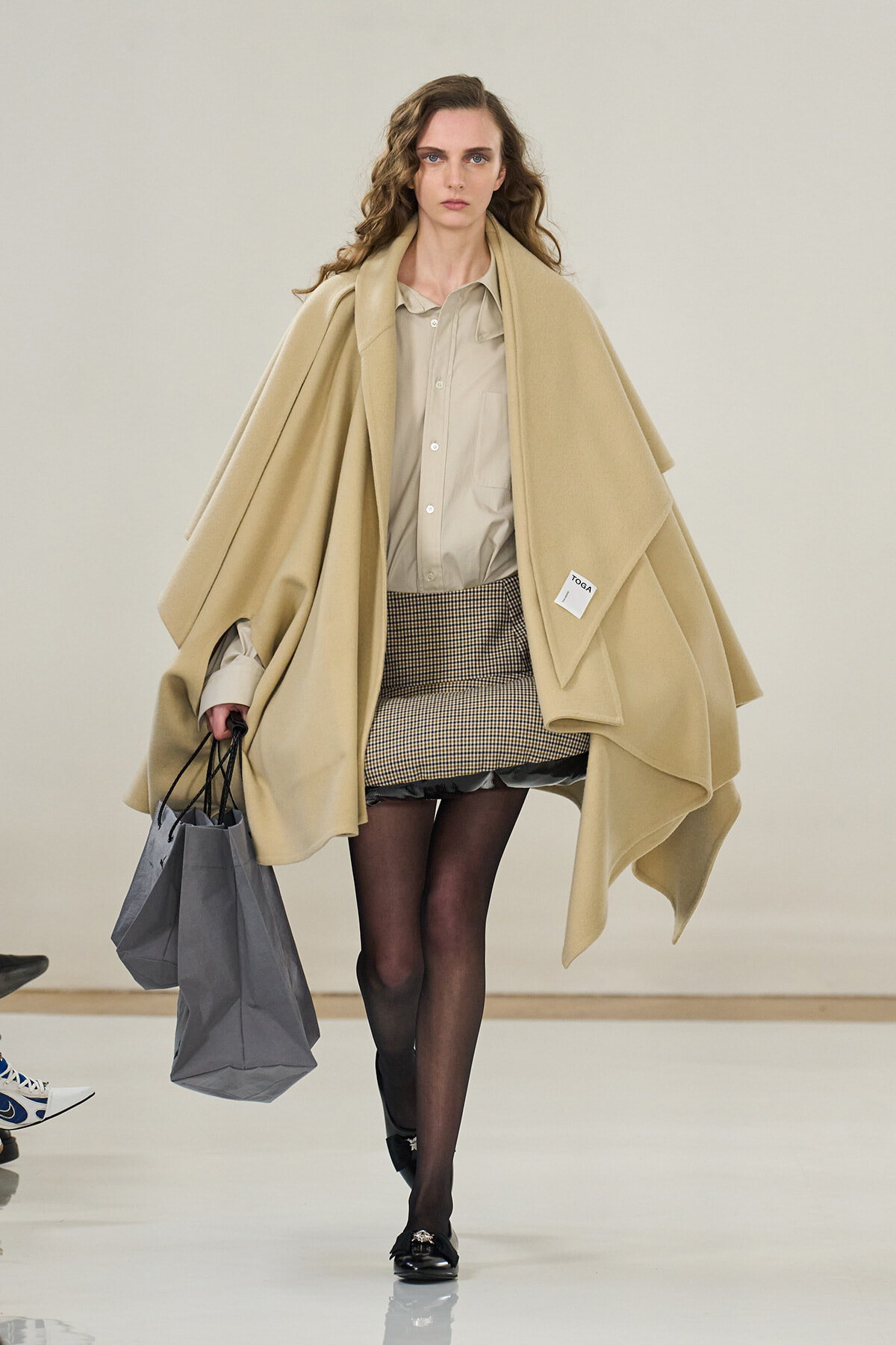 Runway model in a large beige cape over a light shirt and plaid mini skirt, with black tights and loafers, carrying gray shopping bags.