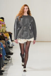 Model on a runway wearing a metallic gray oversized belted top with a white ruffled mini skirt, black tights, and white bow-strap sandals; audience seated along the runway.