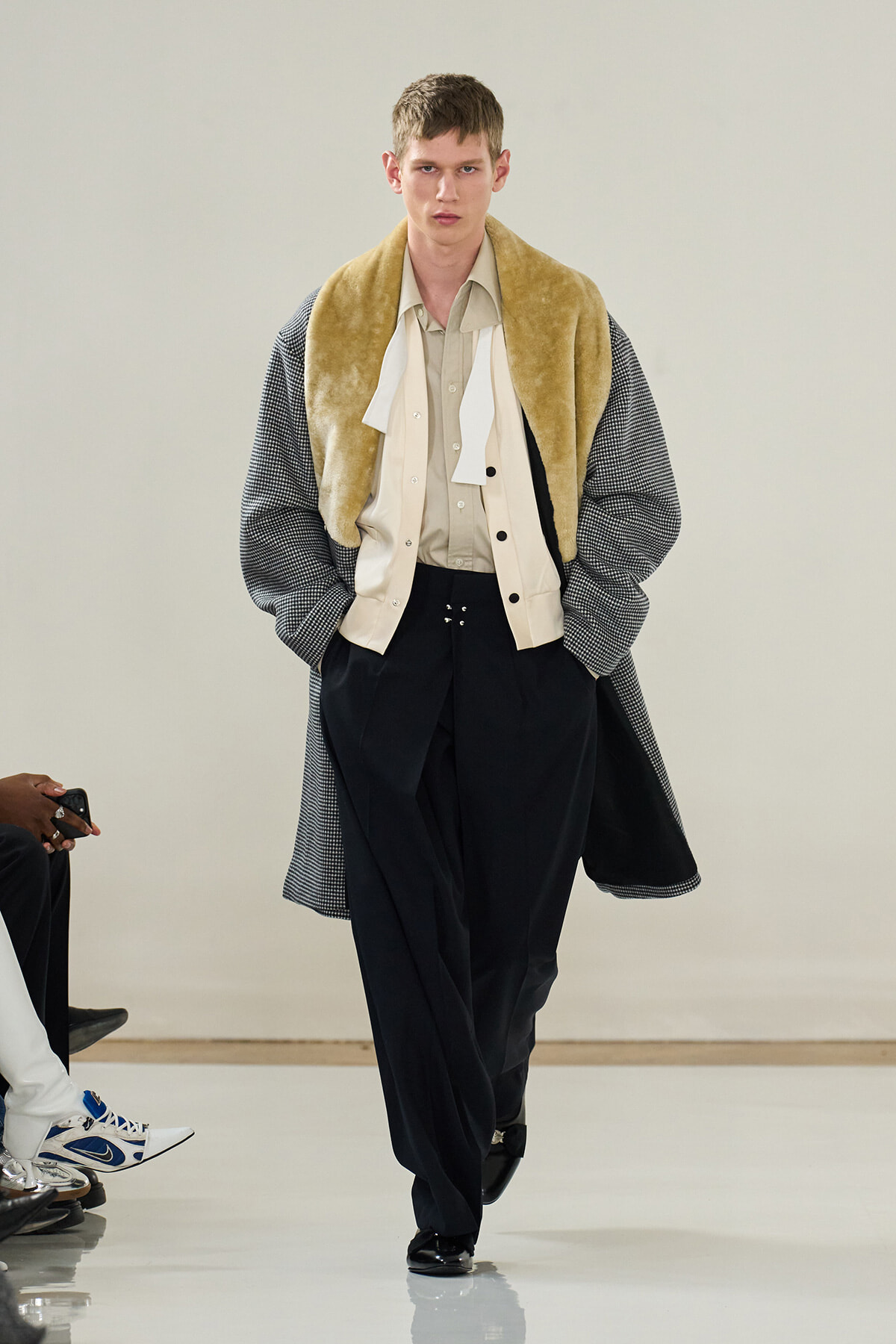 Male model walking a runway in a layered ensemble: beige shirt, cream vest, and a oversized checkered coat draped over the shoulders, with black wide-legged pants and shiny shoes.