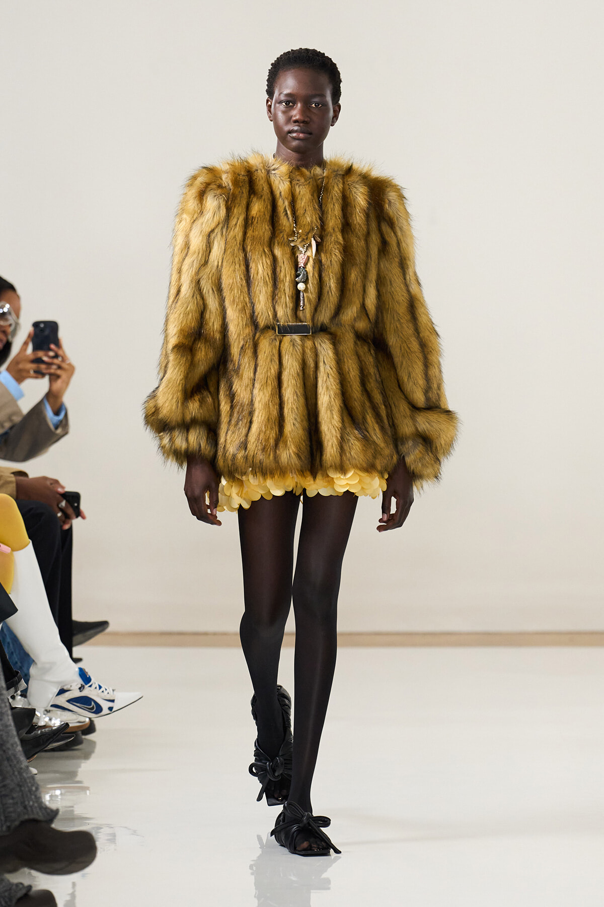 Model walks a fashion runway in a voluminous golden fur coat with a yellow petal-hemged skirt, black tights, and black sandals, accessorized with layered necklaces.