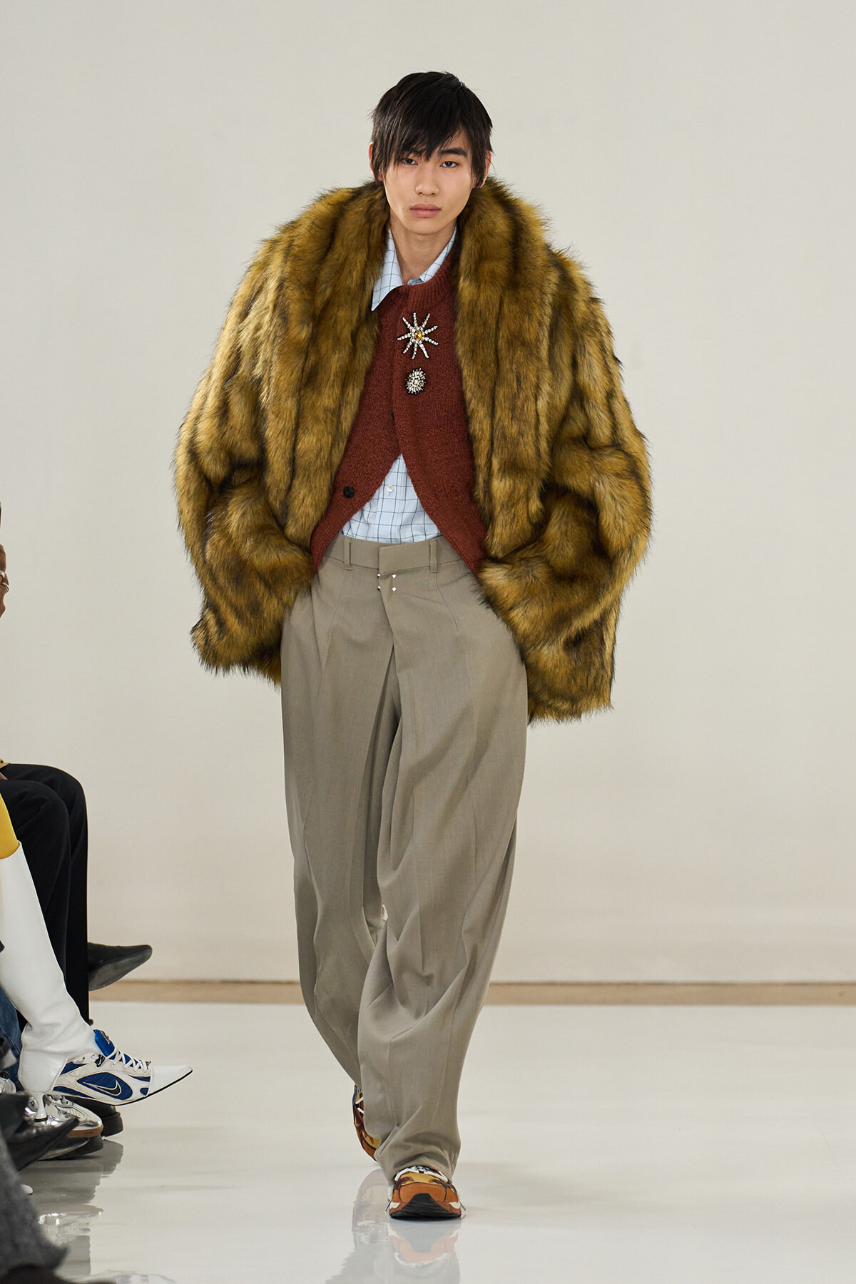 Male runway model in a voluminous brown fur coat over a red cardigan with decorative brooches, paired with a light check shirt and wide beige trousers.