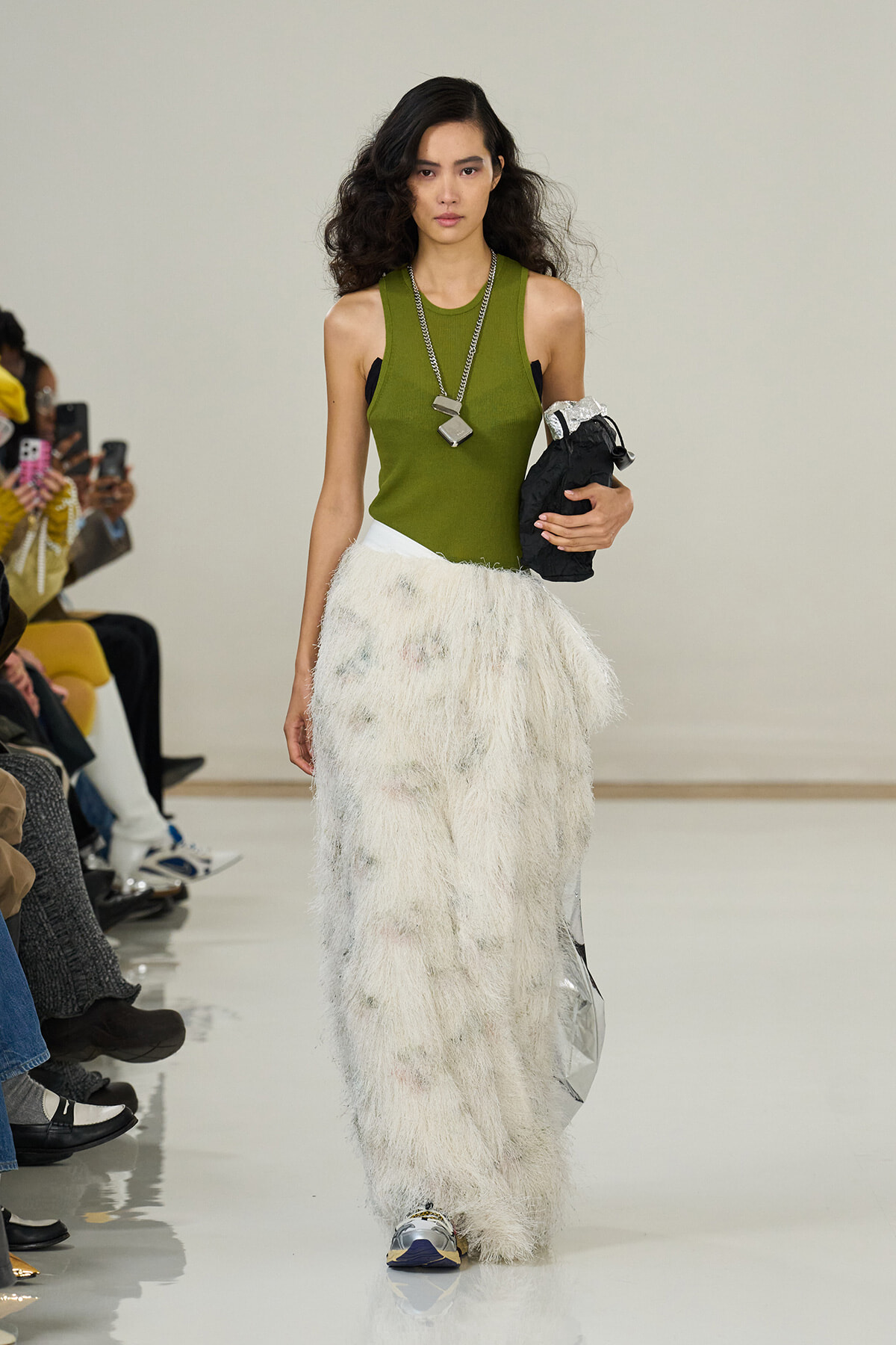 Model walking a runway in a sleeveless olive-green top and a voluminous white shaggy skirt, while holding a black clutch and layered silver necklace. Audience watches from the left.