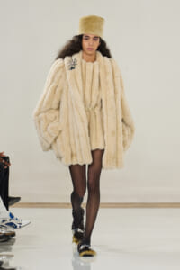 Model walks the runway in a voluminous beige fur coat with a matching fur hat, black tights, and embellished slippers.