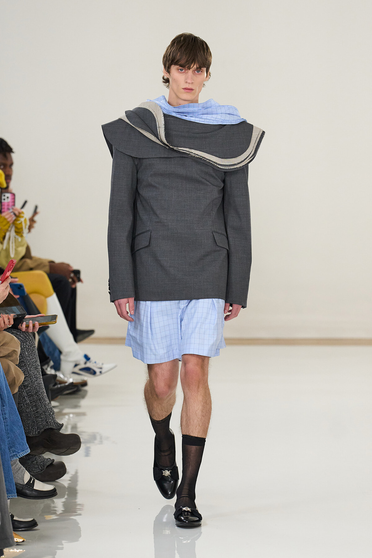 Male model walks a fashion runway in an oversized dark gray coat with a layered sculptural scarf, light blue checked shorts, black socks, and black loafers, while audience watch